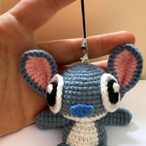 Puede incluir: Mu&ntilde;eco Stitch de ganchillo hecho a mano en tonos azules, blancos y rosas. El mu&ntilde;eco tiene orejas grandes, ojos negros y nariz y pies azules. Est&aacute; sujeto a un cord&oacute;n negro con un cierre plateado.