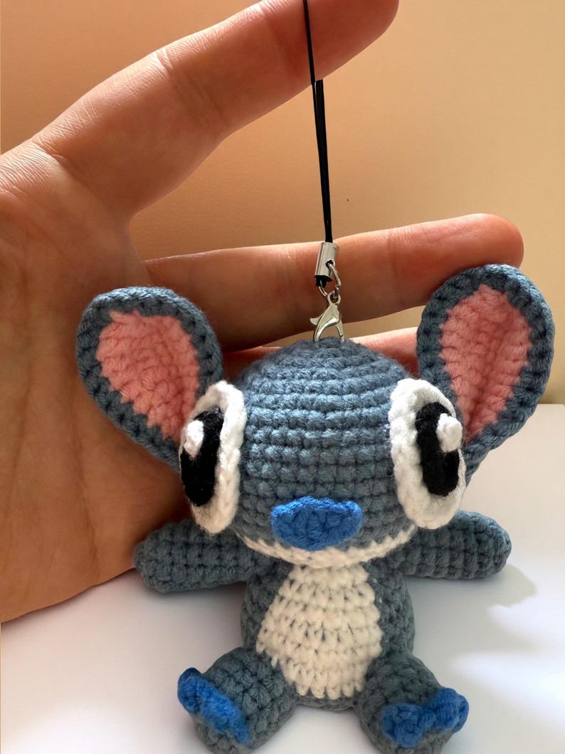 Puede incluir: Mu&ntilde;eco Stitch de ganchillo hecho a mano en tonos azules, blancos y rosas. El mu&ntilde;eco tiene orejas grandes, ojos negros y nariz y pies azules. Est&aacute; sujeto a un cord&oacute;n negro con un cierre plateado.