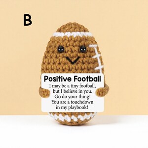May include: A crocheted football with a smiling face and white stripes. The football holds a sign that reads "Positive Football" with an inspirational message. The background is a solid light yellow.