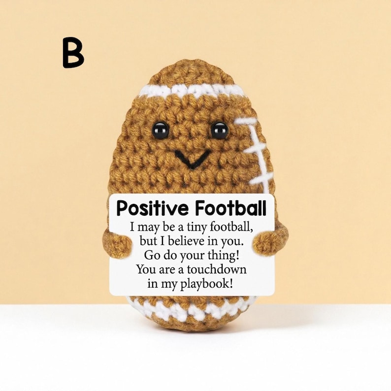 May include: A crocheted football with a smiling face and white stripes. The football holds a sign that reads "Positive Football" with an inspirational message. The background is a solid light yellow.