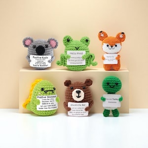 May include: A collection of six crocheted animal figures, including a koala, frog, fox, dinosaur, bear, and turtle. Each figure holds a small sign with positive messages. The koala is gray, the frog is green, the fox is orange, the dinosaur is green, the bear is brown, and the turtle is green.