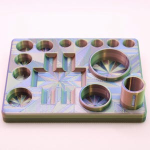 May include: A multi-coloured, geometric-patterned tray with various circular and rectangular cutouts. The tray has a metallic sheen with hues of blue, green, and purple. It features several cylindrical and circular openings of varying sizes.