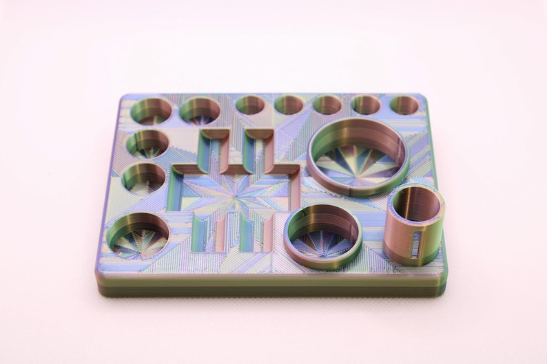 May include: A multi-coloured, geometric-patterned tray with various circular and rectangular cutouts. The tray has a metallic sheen with hues of blue, green, and purple. It features several cylindrical and circular openings of varying sizes.