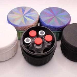 May include: A collection of circular containers in different colours. The containers are white, black, and green with patterned lids. One container is open, showing vials with red caps.