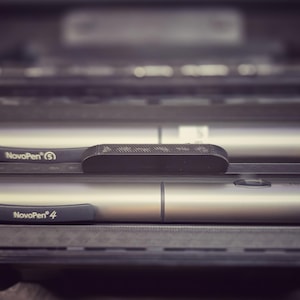 Op de afbeelding: Een zwarte koffer met twee zilveren NovoPen insulinepennen. De pennen zijn cilindrisch met donkergrijze accenten en het NovoPen-logo. Een klein, donkergrijs rechthoekig object ligt tussen de pennen.
