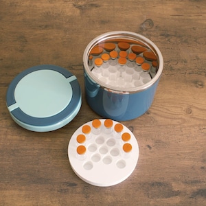 Può includere: Un contenitore isolato blu e argento con coperchio e un vassoio bianco con fiale con tappi arancioni. Il contenitore è aperto, rivelando più fiale all'interno. Il coperchio è azzurro con un bordo blu scuro.