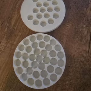 Pode incluir: Duas bandejas circulares brancas com múltiplas reentrâncias redondas. Uma bandeja é parcialmente visível, enquanto a outra é totalmente exibida. As bandejas são feitas de um material sólido e opaco e estão sobre uma superfície de madeira marrom.