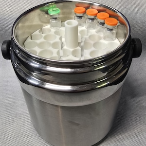Può includere: Un contenitore cilindrico in acciaio inossidabile con due manici laterali neri, contenente un inserto in plastica bianca con molteplici fessure circolari. Diversi piccoli flaconi di vetro con tappi arancioni e verdi sono inseriti nelle fessure.