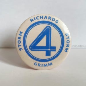 Op de afbeelding: Een ronde, crèmekleurige knoop met een blauwe afbeelding van het nummer 4 in een cirkel. De woorden "RICHARDS", "STORM" en "GRIMM" staan rond de cirkel gedrukt. Een verzamelobject.