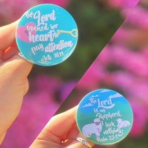Könnte beinhalten: Zwei runde, türkisfarbene Knöpfe mit religiösen Zitaten. Auf einem Knopf steht "The Lord opened her heart to pay attention Acts 16:14." Der andere sagt "The Lord is my Shepherd, I lack nothing. Psalm 23:1." Beide haben weißen Text.