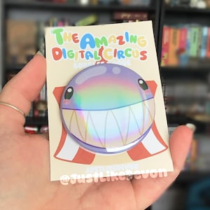 Pode incluir: Um broche circular com acabamento holográfico e iridescente, apresentando um rosto de personagem de desenho animado com olhos grandes e dentes afiados. O broche está montado num cartão de suporte creme com o texto The Amazing Digital Circus.