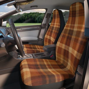 May include: Car seat covers with a plaid pattern in shades of brown, tan, and yellow. The covers are installed in a car, with the steering wheel and dashboard visible. The background shows a view of a green field and trees.