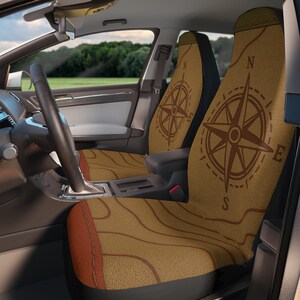 May include: Car seat covers featuring a compass rose design. The covers are tan with a brown compass rose and map-like lines. The car interior includes a steering wheel, dashboard, and a view of a green field and trees.