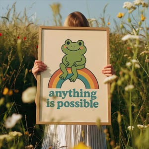 May include: A framed print with a cartoon frog on a rainbow, and the words "anything is possible" in teal. The frame is light brown. The background shows a field of tall grass and flowers, creating a cheerful, nature-inspired scene.