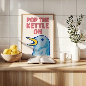 以下が含まれることがあります： 「POP THE KETTLE ON」の文字と、黄色いクチバシを持つ青い鳥のイラストが描かれたフレーム入りのアートプリント。木製の表面に、レモンの入ったボウル、開いた本、マグカップ、緑の入った花瓶が置かれています。
