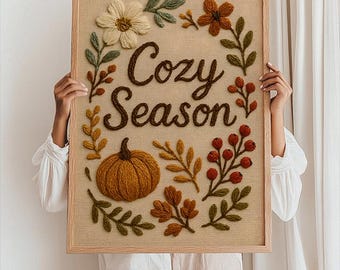 May include: A framed artwork featuring the text "Cozy Season" in brown script, encircled by embroidered autumn motifs. The design incorporates a pumpkin, flowers, leaves, and berries in orange, brown, green, and cream hues. The artwork is presented against a neutral backdrop.