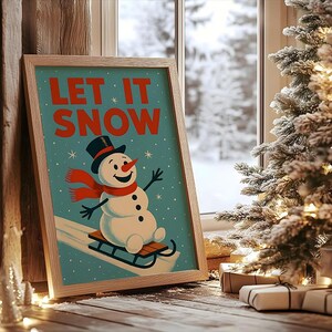 May include: A framed print featuring a snowman on a sled, with a black top hat and red scarf, against a teal backdrop. The words "LET IT SNOW" are in red. The print is in a light wood frame, with a snowy scene and Christmas tree visible in the background.