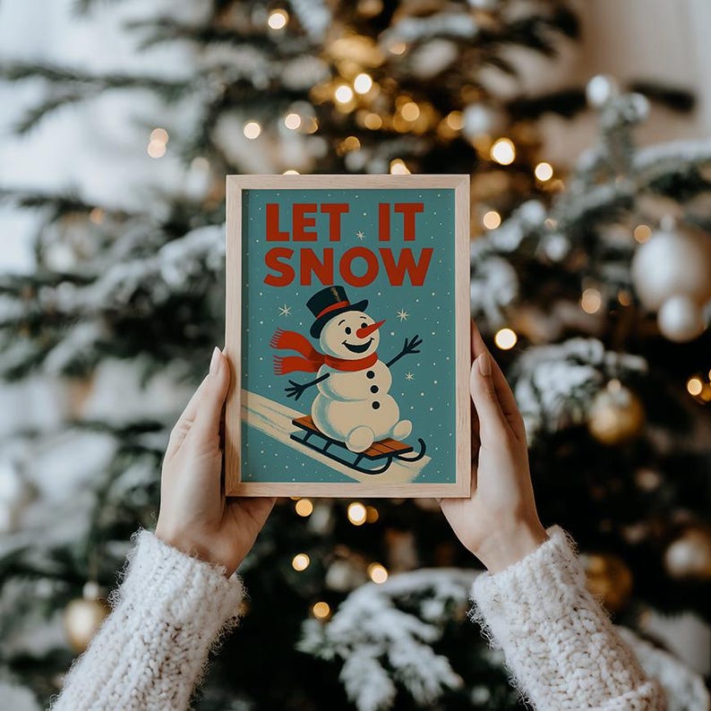 May include: A framed print with the words "LET IT SNOW" in red. The image shows a snowman in a black top hat and red scarf, sledding on a blue background with snowflakes. The frame is made of light-coloured wood.