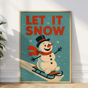May include: A framed winter-themed art print showing a snowman on a sled. The snowman has a black top hat, red scarf, and carrot nose. The words "LET IT SNOW" are in red against a teal background.