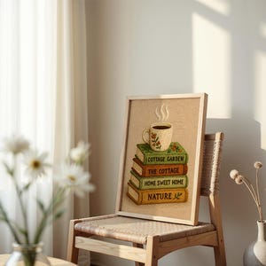 May include: A framed picture showing a stack of books with the titles "Cottage Garden", "The Cottage", "Home Sweet Home", and "Nature". A cup of coffee sits on top. The artwork is displayed on a wooden chair.