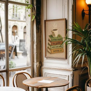 May include: A framed picture shows a coffee cup on a stack of books, with titles including "Cottage Garden" and "Home Sweet Home." The artwork hangs in a cafe, with a wooden table and a window overlooking a street scene. The overall aesthetic is warm and inviting.