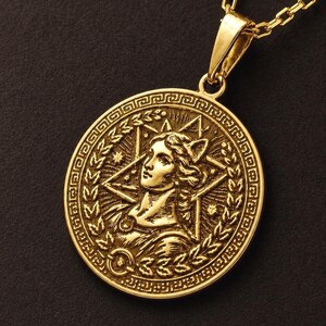 May include: A gold-toned circular pendant necklace featuring a relief portrait of a figure in profile, surrounded by a laurel wreath and a star-shaped geometric pattern. The pendant is suspended from a matching gold-toned chain link necklace.