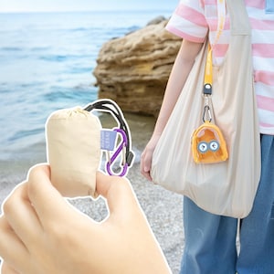 Op de afbeelding: Een beige trekkoordtas met een zwart koord en een paarse karabijnhaak. Een kleine gele sleutelhanger in de vorm van een rugzak met een cartoongezicht hangt aan een gele lanyard die aan een beige draagtas is bevestigd. De scène speelt zich af op het strand.