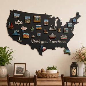 May include: A black wall-mounted map of the United States featuring the text "With you, I am home" in white script. The map is decorated with various colorful souvenir magnets depicting landmarks, cities, and states across the country.