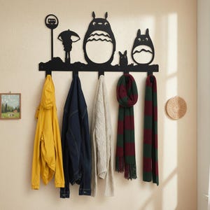 May include: A black metal wall-mounted coat rack featuring a silhouette design of characters from a popular animated film. The rack holds a yellow jacket, denim jacket, cream sweater, and two striped scarves. A small framed picture and a woven wall hanging are also visible.