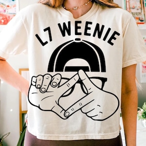 Può includere: T-shirt color crema con un design grafico nero. Il design include il testo "L7 WEENIE" sopra un berretto da baseball nero, occhiali e due mani che fanno un gesto. La maglietta è uno stile casual.