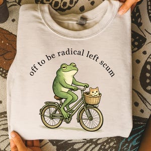 May include: A light gray sweatshirt with the text "off to be radical left scum." The graphic shows a green frog riding a bicycle with a small cat in a basket. The bicycle is light green.