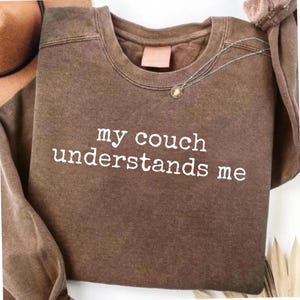 May include: A brown crewneck sweatshirt with a vintage wash finish. The front features white text that reads, "my couch understands me" in a typewriter-style font. The garment has long sleeves, a ribbed neckline, and a relaxed fit design.