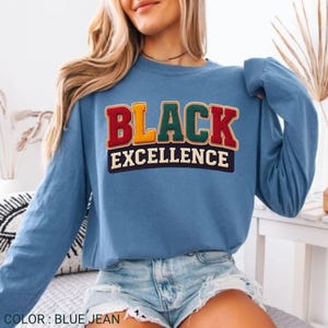 Op de afbeelding: Een blauw spijkerstof sweatshirt met lange mouwen met de tekst "BLACK EXCELLENCE" in een kleurrijk, geborduurd ontwerp. De sweatshirt heeft een effen blauwe kleur en een ronde hals.