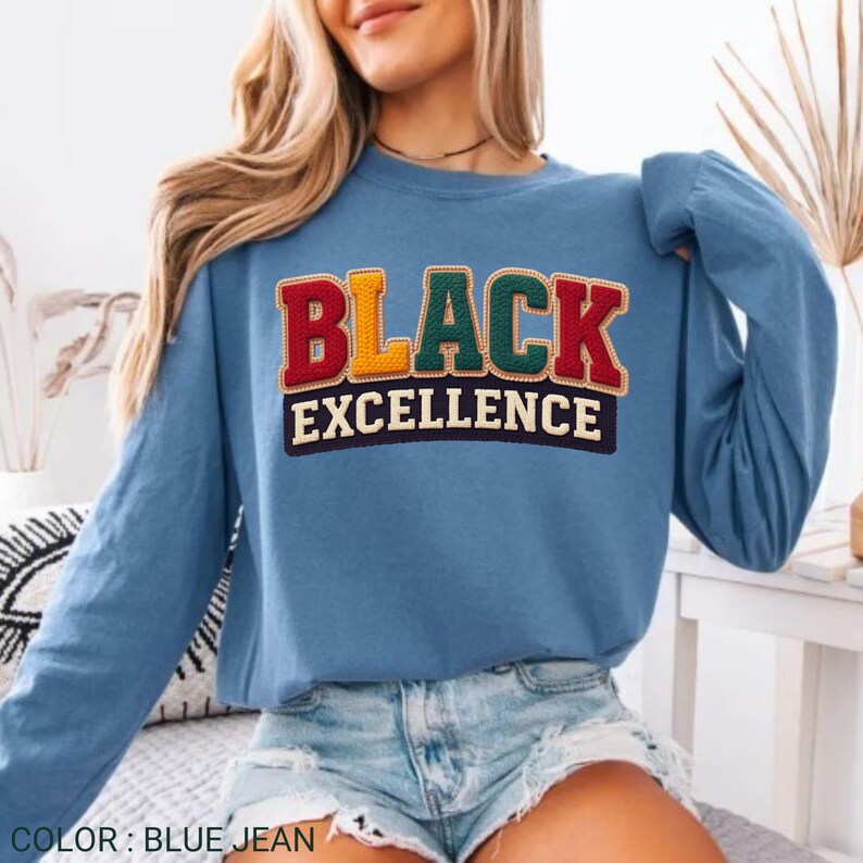 Op de afbeelding: Een blauw spijkerstof sweatshirt met lange mouwen met de tekst "BLACK EXCELLENCE" in een kleurrijk, geborduurd ontwerp. De sweatshirt heeft een effen blauwe kleur en een ronde hals.