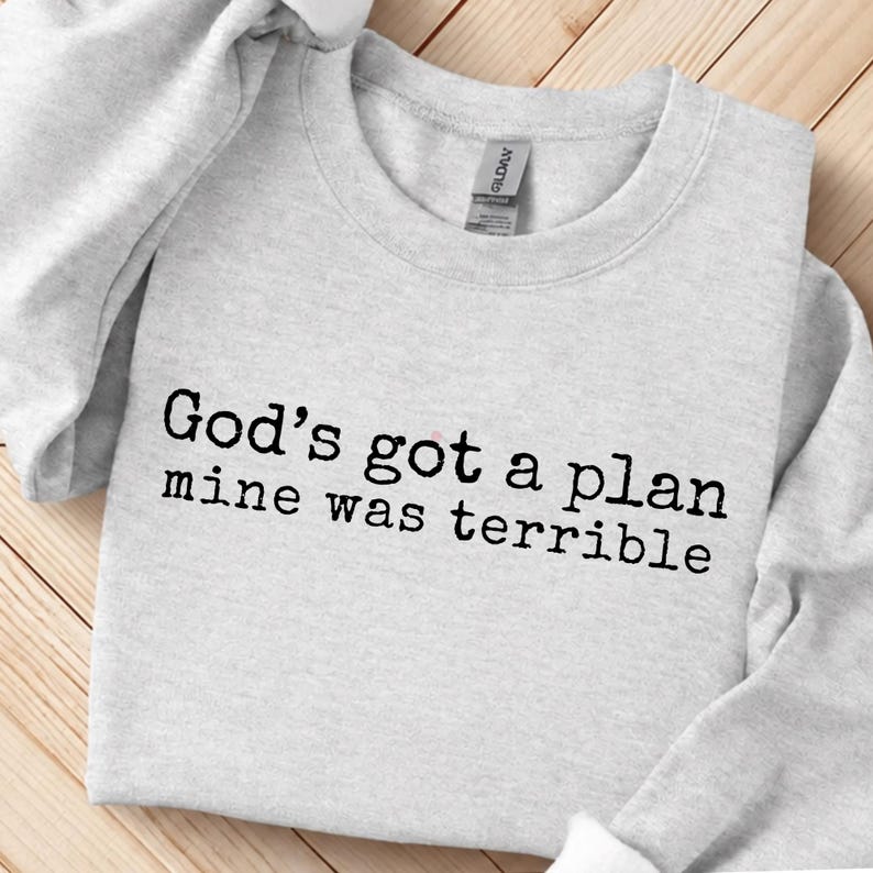 May include: A light grey crewneck sweatshirt featuring the black text "God's got a plan mine was terrible" printed across the chest. The garment has long sleeves, a ribbed neckline, and a Gildan brand label visible inside the collar area.