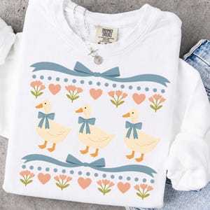 Puede incluir: Una sudadera blanca de manga larga con cuello redondo que presenta un estampado gr&aacute;fico de tres patos de color crema con lazos azules. El dise&ntilde;o incluye filas de cintas azules, peque&ntilde;as flores rosas y corazones color melocot&oacute;n. La etiqueta dice Comfort Colors.