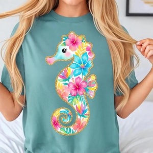 May include: A teal t-shirt featuring a colorful seahorse design. The seahorse is adorned with pink, blue, and yellow floral patterns, outlined with gold glitter. The design is centered on the shirt, creating a vibrant and eye-catching look.