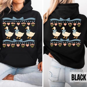 Puede incluir: Una sudadera negra con capucha que presenta un estampado gr&aacute;fico de tres patos de color crema con lazos azules. El dise&ntilde;o incluye filas de cintas azul claro, corazones rosas y peque&ntilde;as flores verdes dispuestas en un patr&oacute;n horizontal.