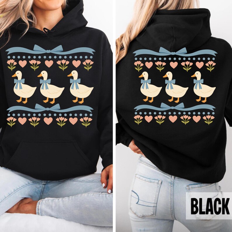 Puede incluir: Una sudadera negra con capucha que presenta un estampado gr&aacute;fico de tres patos de color crema con lazos azules. El dise&ntilde;o incluye filas de cintas azul claro, corazones rosas y peque&ntilde;as flores verdes dispuestas en un patr&oacute;n horizontal.