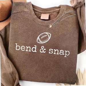 Pode incluir: Uma camisola castanha de gola redonda com um gráfico branco de uma bola de futebol americano sobre o texto bend & snap. A peça tem um acabamento lavado de estilo vintage com mangas compridas, uma gola canelada e uma etiqueta visível.