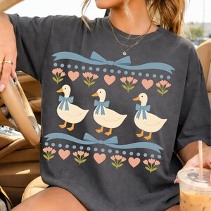 Puede incluir: Una camiseta gris carb&oacute;n con un estampado gr&aacute;fico de tres patos de color crema con lazos azules. El dise&ntilde;o incluye filas de cintas azul claro, flores rosas y peque&ntilde;os corazones rosas dispuestos en un patr&oacute;n horizontal repetitivo.