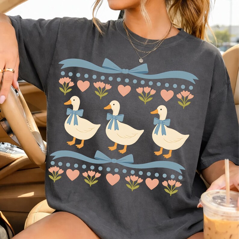 Puede incluir: Una camiseta gris carb&oacute;n con un estampado gr&aacute;fico de tres patos de color crema con lazos azules. El dise&ntilde;o incluye filas de cintas azul claro, flores rosas y peque&ntilde;os corazones rosas dispuestos en un patr&oacute;n horizontal repetitivo.