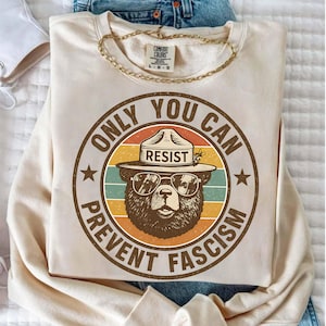 May include: Cream-colored sweatshirt with a circular graphic featuring a bear wearing sunglasses and a ranger hat. The graphic includes the text "ONLY YOU CAN PREVENT FASCISM" in a vintage style. A gold chain necklace is draped on the sweatshirt.