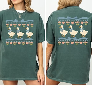 Puede incluir: Una camiseta verde oscuro con un estampado gr&aacute;fico repetitivo de tres patos blancos con lazos azules, dispuestos en fila entre hileras horizontales de cintas azul claro, corazones rosas y peque&ntilde;as flores rosas en el frente y la espalda.