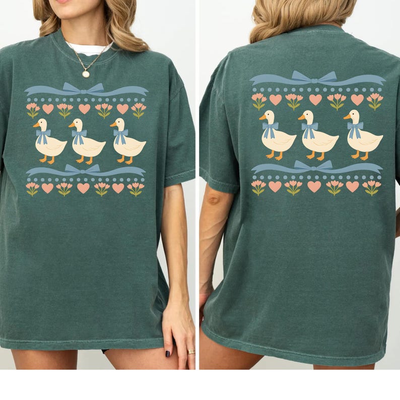 Puede incluir: Una camiseta verde oscuro con un estampado gr&aacute;fico repetitivo de tres patos blancos con lazos azules, dispuestos en fila entre hileras horizontales de cintas azul claro, corazones rosas y peque&ntilde;as flores rosas en el frente y la espalda.