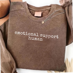 Può includere: Una felpa girocollo marrone con finitura lavata vintage, caratterizzata dalla scritta bianca "emotional support human" stampata sul petto. Il capo presenta maniche lunghe, un colletto a coste e un aspetto del tessuto morbido e strutturato.