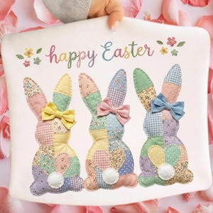 Peut inclure: Un coussin blanc sur le thème de Pâques avec trois lapins en patchwork avec des nœuds colorés et des motifs floraux. Les mots "happy Easter" sont brodés en couleurs pastel au-dessus des lapins.