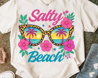 May include: Cream-coloured t-shirt with the words "Salty Beach" in pink and teal. The design features sunglasses with a leopard print frame, and a beach scene reflected in the lenses. Pink flowers and green leaves surround the sunglasses.