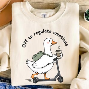 May include: Cream-colored sweatshirt with a graphic of a white goose riding a scooter, carrying a backpack and a coffee cup. The text "Off to regulate emotions" is printed above the goose.