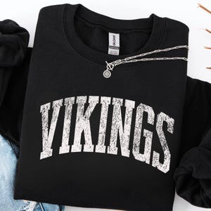 Op de afbeelding: Een zwart sweatshirt met lange mouwen en ronde hals met het woord VIKINGS in een groot, gebogen, verweerd wit lettertype op de borst gedrukt. Het kledingstuk heeft een Gildan-label bij de binnenhals en een zilverkleurige ketting.